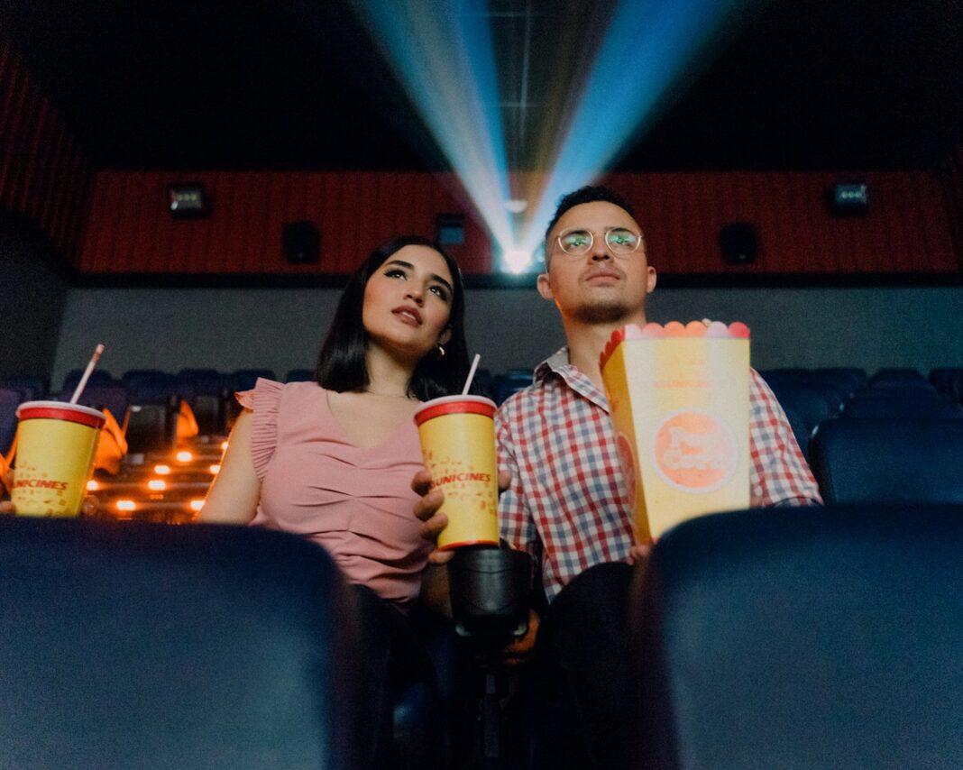 2 people sitting on blue leather chair at the movies holding cup and pop corns